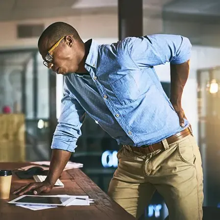 Person leaning on a desk with one hand, holding lower back with the other, papers and a coffee cup on the table.